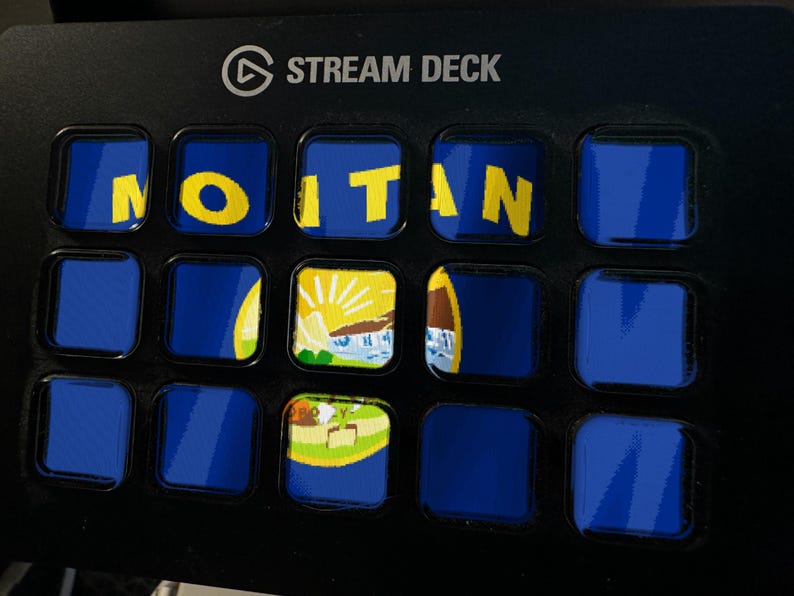 Puede incluir: Un controlador Stream Deck negro con una cuadr&iacute;cula de quince botones cuadrados. Los botones muestran un fondo azul con el texto amarillo MONTANA y un gr&aacute;fico parcial del sello estatal, con varios botones mostrando solo azul.