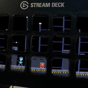 May include: A black Stream Deck device featuring fifteen square LCD keys arranged in a grid. The keys display a pixelated video game scene with a blue character, a red block, and industrial platforms against a dark background.