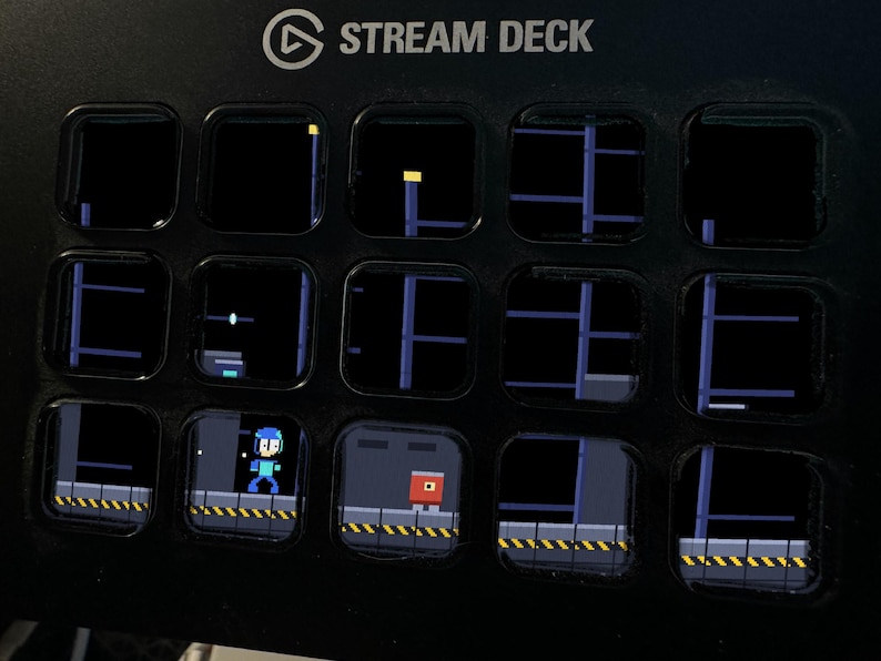 May include: A black Stream Deck device featuring fifteen square LCD keys arranged in a grid. The keys display a pixelated video game scene with a blue character, a red block, and industrial platforms against a dark background.
