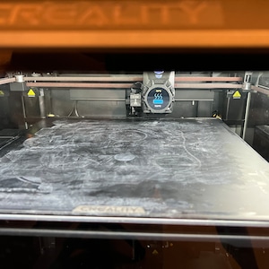 Op de afbeelding: Een zwarte 3D-printer met een glazen printbed en een digitaal display dat "SSS" toont.