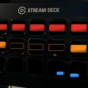 May include: A black Stream Deck control console featuring a grid of fifteen square buttons. The top row displays five solid red rectangular icons. The middle row shows two solid orange icons and three orange outlines. The bottom row has two blue icons.