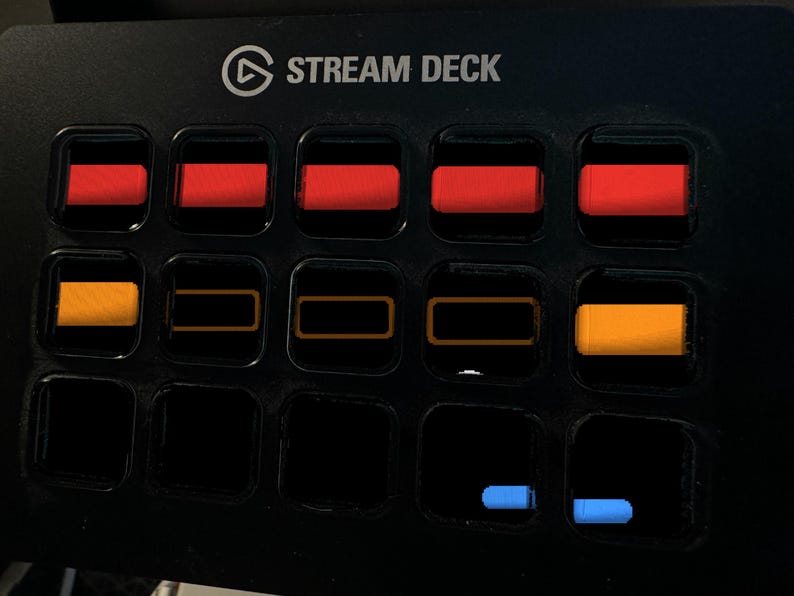 May include: A black Stream Deck control console featuring a grid of fifteen square buttons. The top row displays five solid red rectangular icons. The middle row shows two solid orange icons and three orange outlines. The bottom row has two blue icons.