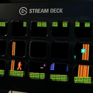 May include: A black Stream Deck controller featuring fifteen square LCD keys. Each key displays a pixelated graphic from a retro video game, including green platforms, orange vertical pillars, and a small orange character figure on a black background.