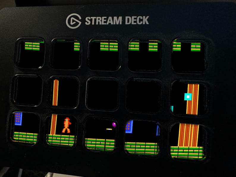 May include: A black Stream Deck controller featuring fifteen square LCD keys. Each key displays a pixelated graphic from a retro video game, including green platforms, orange vertical pillars, and a small orange character figure on a black background.