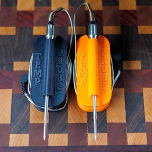 May include: Two temperature probes, one black and one orange, with the words "TEMP PROBE" embossed on the front. Each probe has a stainless steel tip and a silver cable. The probes are resting on a wooden cutting board.