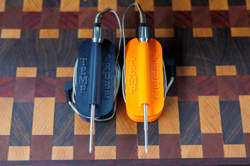 May include: Two temperature probes, one black and one orange, with the words "TEMP PROBE" embossed on the front. Each probe has a stainless steel tip and a silver cable. The probes are resting on a wooden cutting board.