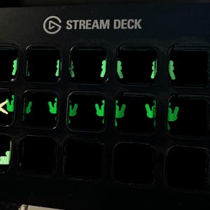 May include: A black Stream Deck control console featuring a grid of fifteen square buttons. The buttons display illuminated green icons, including a yellow asterisk symbol on the second row and a green staircase graphic on the bottom left corner.