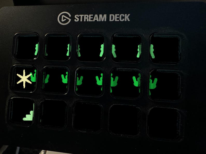 May include: A black Stream Deck control console featuring a grid of fifteen square buttons. The buttons display illuminated green icons, including a yellow asterisk symbol on the second row and a green staircase graphic on the bottom left corner.