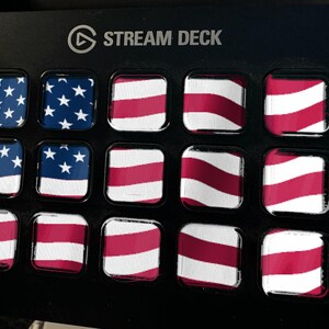 May include: A black Stream Deck device featuring fifteen square keys arranged in a grid. Each key displays a portion of the American flag, with the blue field and white stars on the left and red and white horizontal stripes across the remaining keys.