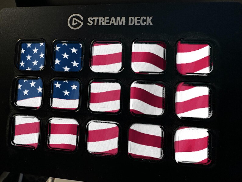 May include: A black Stream Deck device featuring fifteen square keys arranged in a grid. Each key displays a portion of the American flag, with the blue field and white stars on the left and red and white horizontal stripes across the remaining keys.