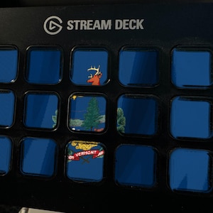 Może przedstawiać: Czarna konsola sterująca Stream Deck z piętnastoma kwadratowymi przyciskami. Przyciski wyświetlają siatkę niebieskich ekran&oacute;w, przy czym trzy konkretne przyciski pokazują głowę jelenia, sosnę oraz grafikę z napisem Vermont.