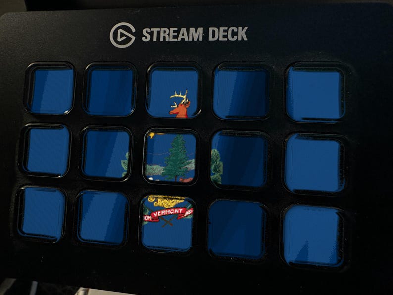 Może przedstawiać: Czarna konsola sterująca Stream Deck z piętnastoma kwadratowymi przyciskami. Przyciski wyświetlają siatkę niebieskich ekran&oacute;w, przy czym trzy konkretne przyciski pokazują głowę jelenia, sosnę oraz grafikę z napisem Vermont.