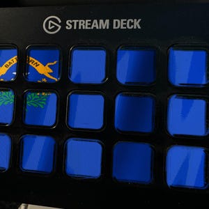 Pode incluir: Uma consola de controlo Stream Deck preta com quinze teclas quadradas dispostas numa grelha. As duas teclas superiores esquerdas mostram parte da bandeira do estado do Nevada com texto amarelo e um gr&aacute;fico verde de artem&iacute;sia. As restantes treze s&atilde;o azuis.