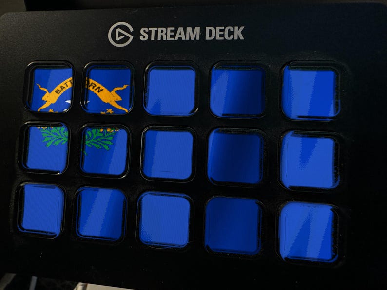 Pode incluir: Uma consola de controlo Stream Deck preta com quinze teclas quadradas dispostas numa grelha. As duas teclas superiores esquerdas mostram parte da bandeira do estado do Nevada com texto amarelo e um gr&aacute;fico verde de artem&iacute;sia. As restantes treze s&atilde;o azuis.