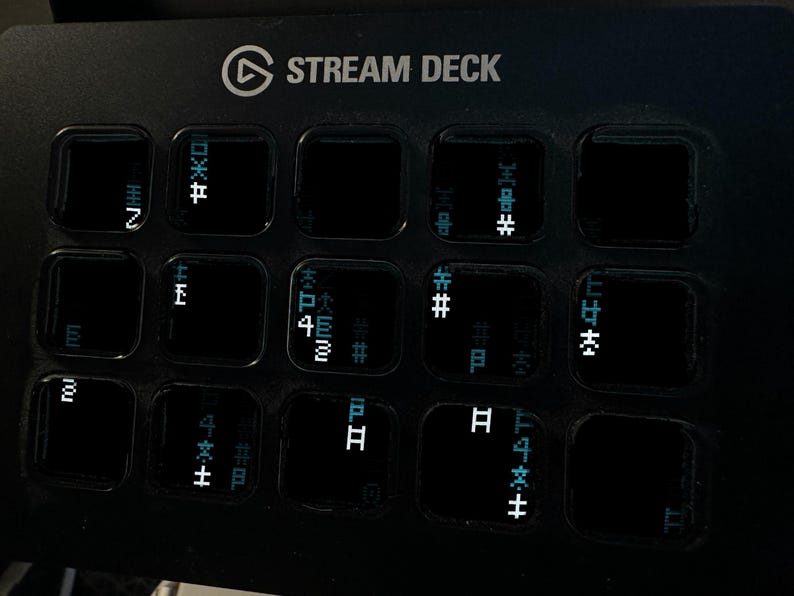 Op de afbeelding: Een zwarte Stream Deck-bedieningsconsole met een raster van vijftien vierkante knoppen. Elke knop toont een unieke, glitchy digitale afbeelding bestaande uit witte en lichtblauwe gepixelde symbolen, tekens en abstracte geometrische vormen.