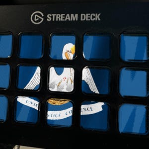 Pode incluir: Um controlador Stream Deck preto com quinze bot&otilde;es quadrados dispostos em uma grade. Os bot&otilde;es exibem uma imagem fragmentada da bandeira do estado da Louisiana, mostrando um pelicano branco, um ninho e uma faixa com o texto Union Justice Confidence.