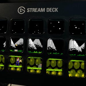 Peut inclure: Un appareil Stream Deck noir dot&eacute; de quinze touches LCD carr&eacute;es dispos&eacute;es en grille. La rang&eacute;e sup&eacute;rieure affiche un ciel nocturne &eacute;toil&eacute;, la rang&eacute;e centrale montre des sommets montagneux pix&eacute;lis&eacute;s et la rang&eacute;e inf&eacute;rieure un personnage.