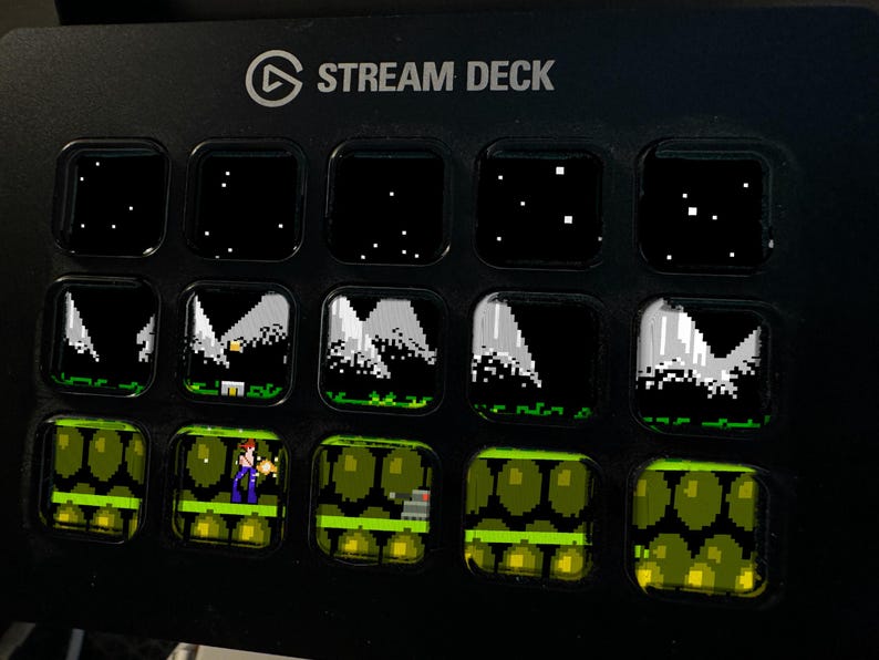 Peut inclure: Un appareil Stream Deck noir dot&eacute; de quinze touches LCD carr&eacute;es dispos&eacute;es en grille. La rang&eacute;e sup&eacute;rieure affiche un ciel nocturne &eacute;toil&eacute;, la rang&eacute;e centrale montre des sommets montagneux pix&eacute;lis&eacute;s et la rang&eacute;e inf&eacute;rieure un personnage.