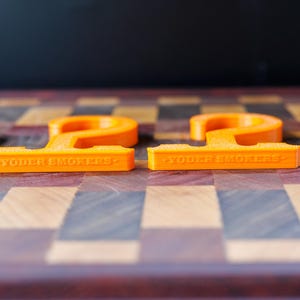 May include: Two bright orange, stylized letter "P" shaped objects with the text "YODER SMOKERS" on the base. The objects are on a wooden checkerboard surface.