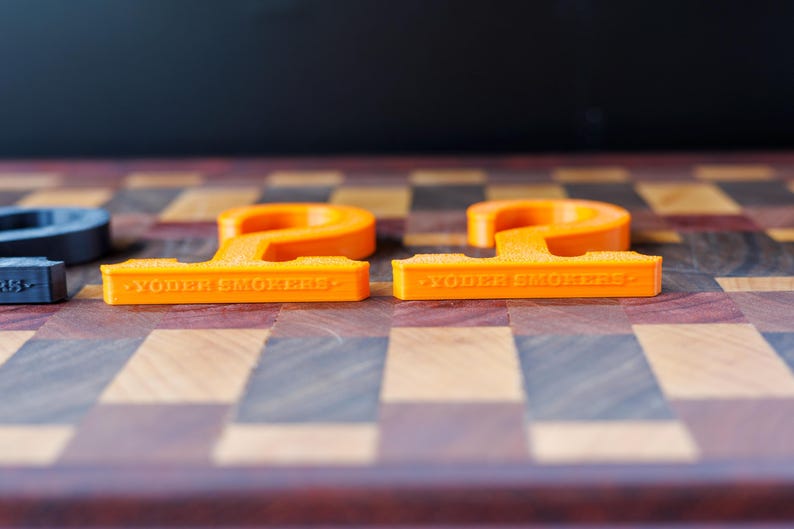 May include: Two bright orange, stylized letter "P" shaped objects with the text "YODER SMOKERS" on the base. The objects are on a wooden checkerboard surface.