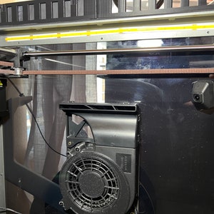Op de afbeelding: Close-up van de interne werking van een 3D-printer, met een bruine riem, een zilveren staaf en een zwarte ventilator met een beschermkap. De printer heeft een zwart en zilveren frame.