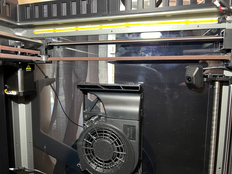 Op de afbeelding: Close-up van de interne werking van een 3D-printer, met een bruine riem, een zilveren staaf en een zwarte ventilator met een beschermkap. De printer heeft een zwart en zilveren frame.