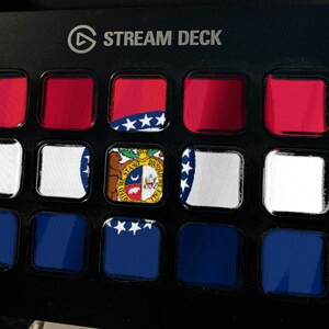 K&ouml;nnte beinhalten: Ein schwarzer Stream Deck-Controller mit einem Raster aus f&uuml;nfzehn quadratischen Tasten. Die Tasten zeigen eine fragmentierte Grafik der Flagge des Bundesstaates Missouri mit roten, wei&szlig;en und blauen horizontalen Streifen, einem zentralen Siegel und wei&szlig;en Sternen.