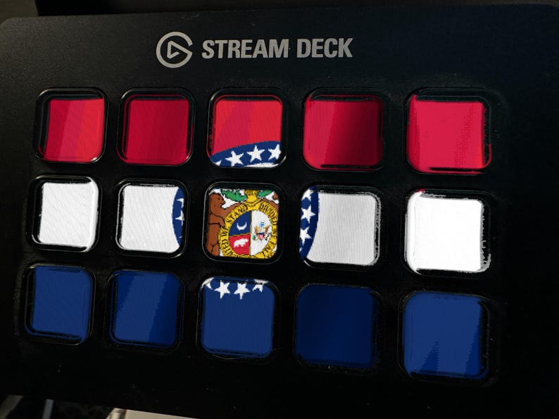 K&ouml;nnte beinhalten: Ein schwarzer Stream Deck-Controller mit einem Raster aus f&uuml;nfzehn quadratischen Tasten. Die Tasten zeigen eine fragmentierte Grafik der Flagge des Bundesstaates Missouri mit roten, wei&szlig;en und blauen horizontalen Streifen, einem zentralen Siegel und wei&szlig;en Sternen.
