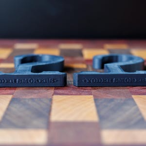 May include: Two dark gray, number 2 shaped objects with the text "WOODEN SMOKERS" on the bottom, resting on a wooden checkerboard. The objects are 3D printed and have a matte finish. Orange game pieces are visible in the background.