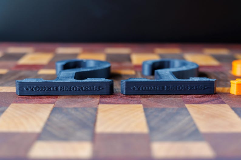 May include: Two dark gray, number 2 shaped objects with the text "WOODEN SMOKERS" on the bottom, resting on a wooden checkerboard. The objects are 3D printed and have a matte finish. Orange game pieces are visible in the background.