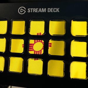 Pode incluir: Um controlador Stream Deck preto com quinze teclas quadradas dispostas em grelha. As teclas apresentam um fundo amarelo com um s&iacute;mbolo solar vermelho Zia da bandeira do estado do Novo M&eacute;xico que se estende pelo centro do layout.