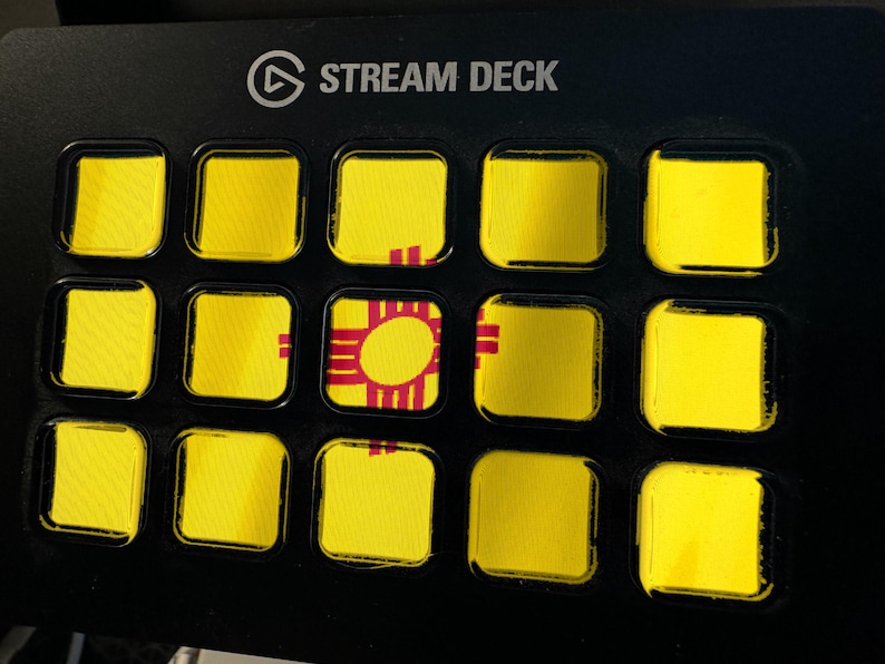 Pode incluir: Um controlador Stream Deck preto com quinze teclas quadradas dispostas em grelha. As teclas apresentam um fundo amarelo com um s&iacute;mbolo solar vermelho Zia da bandeira do estado do Novo M&eacute;xico que se estende pelo centro do layout.