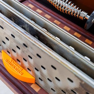 Op de afbeelding: Close-up van de binnenkant van een smoker, met geperforeerde metalen rekken en een oranje wigvormig stuk met de tekst "YODER SMOKERS". De binnenkant van de smoker staat op een houten oppervlak.