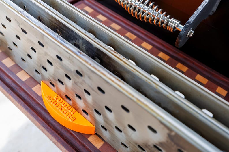 Op de afbeelding: Close-up van de binnenkant van een smoker, met geperforeerde metalen rekken en een oranje wigvormig stuk met de tekst "YODER SMOKERS". De binnenkant van de smoker staat op een houten oppervlak.