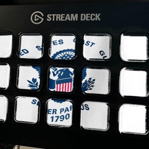Op de afbeelding: Een zwart Stream Deck bedieningspaneel met vijftien vierkante knoppen. Verschillende knoppen tonen delen van het embleem en de tekst van de United States Coast Guard, waaronder 'United States Coast Guard' en 'Semper Paratus 1790', terwijl andere leeg zijn.