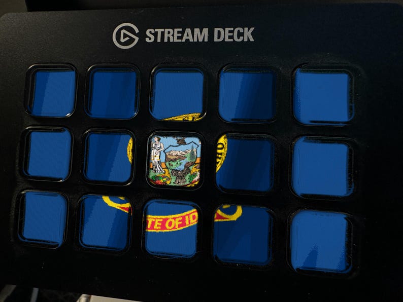 K&ouml;nnte beinhalten: Ein schwarzer Stream Deck-Controller mit einem Raster aus f&uuml;nfzehn quadratischen Tasten. Die Tasten zeigen ein fragmentiertes Bild der Flagge des US-Bundesstaates Idaho, wobei die mittlere Taste das Staatssiegel und die untere Reihe den Text STATE OF IDAHO zeigt.