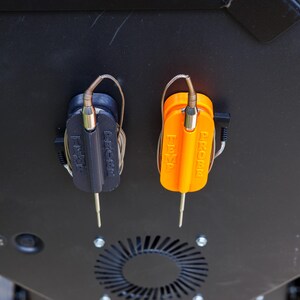May include: Close-up of a black and orange grill temperature probe set. The probes are attached to a black surface. The orange probe has the word "PROBE" printed on it. The black probe has the word "P-CHEF" printed on it.