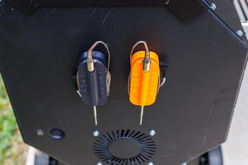 May include: Close-up of a black and orange grill temperature probe set. The probes are attached to a black surface. The orange probe has the word "PROBE" printed on it. The black probe has the word "P-CHEF" printed on it.