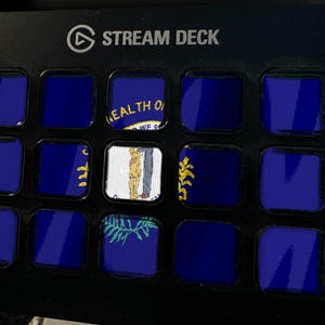 K&ouml;nnte beinhalten: Ein schwarzer Stream Deck-Controller mit einem Raster aus f&uuml;nfzehn quadratischen Tasten. Jede Taste zeigt einen Teil der Flagge des Bundesstaates Nebraska, die durch einen tiefblauen Hintergrund und ein zentrales Siegel mit Schmied und Weizen gekennzeichnet ist.