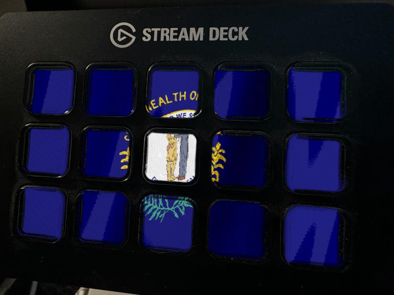 K&ouml;nnte beinhalten: Ein schwarzer Stream Deck-Controller mit einem Raster aus f&uuml;nfzehn quadratischen Tasten. Jede Taste zeigt einen Teil der Flagge des Bundesstaates Nebraska, die durch einen tiefblauen Hintergrund und ein zentrales Siegel mit Schmied und Weizen gekennzeichnet ist.