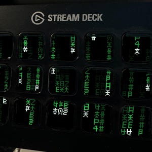 May include: A black Stream Deck control console featuring a grid of fifteen square keys. Each key displays a glowing green digital matrix pattern with various symbols and characters, resembling falling code, set against a dark background.