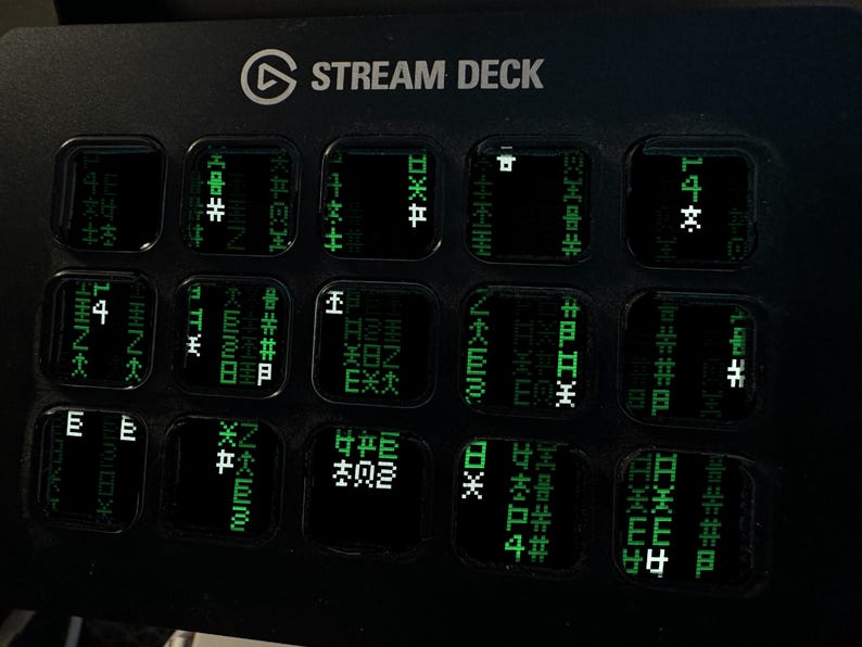 May include: A black Stream Deck control console featuring a grid of fifteen square keys. Each key displays a glowing green digital matrix pattern with various symbols and characters, resembling falling code, set against a dark background.