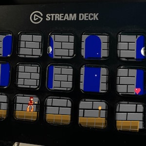 May include: A black Stream Deck device featuring fifteen square LCD keys arranged in a grid. Each key displays a portion of a pixelated video game scene, including gray brick walls, blue doorways, a small character, a torch, and a red heart icon.