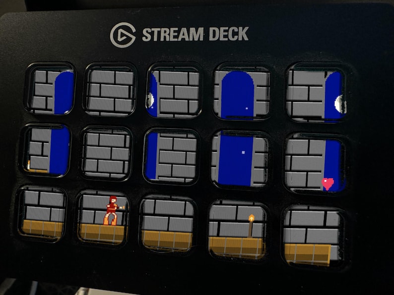 May include: A black Stream Deck device featuring fifteen square LCD keys arranged in a grid. Each key displays a portion of a pixelated video game scene, including gray brick walls, blue doorways, a small character, a torch, and a red heart icon.