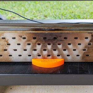 Op de afbeelding: Een rechthoekige rokerbox van roestvrij staal met meerdere gaten, rustend op een zwart oppervlak. Een oranje, gebogen stuk met de tekst "YODER MONSTER" staat voor de box. De achtergrond toont groen gras.