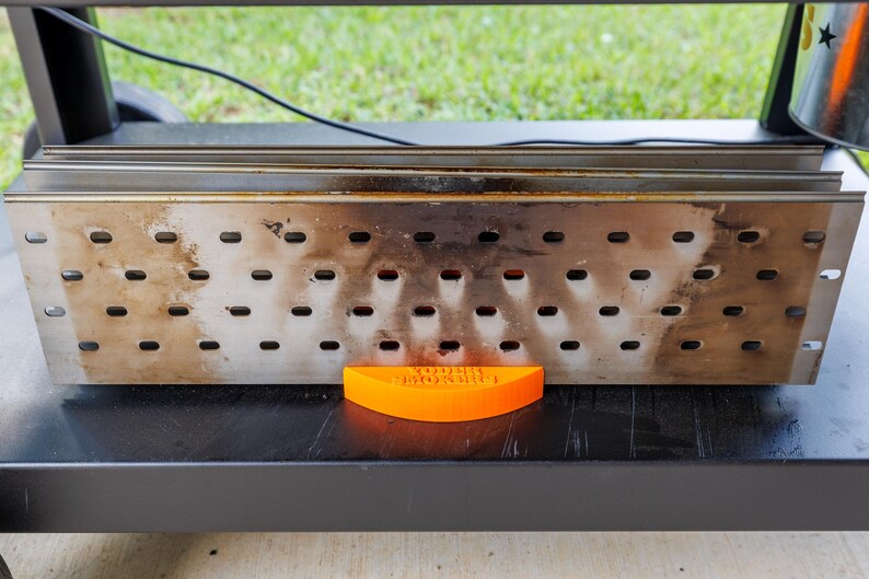 Op de afbeelding: Een rechthoekige rokerbox van roestvrij staal met meerdere gaten, rustend op een zwart oppervlak. Een oranje, gebogen stuk met de tekst "YODER MONSTER" staat voor de box. De achtergrond toont groen gras.