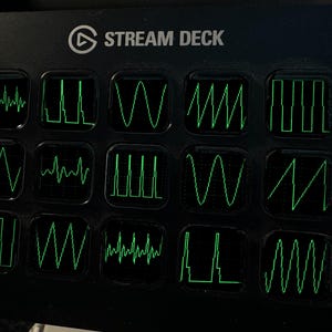 May include: A black Stream Deck controller featuring fifteen square keys arranged in a grid. Each key displays a unique, glowing green waveform graphic, including sine, square, triangle, and sawtooth waves against a dark background.