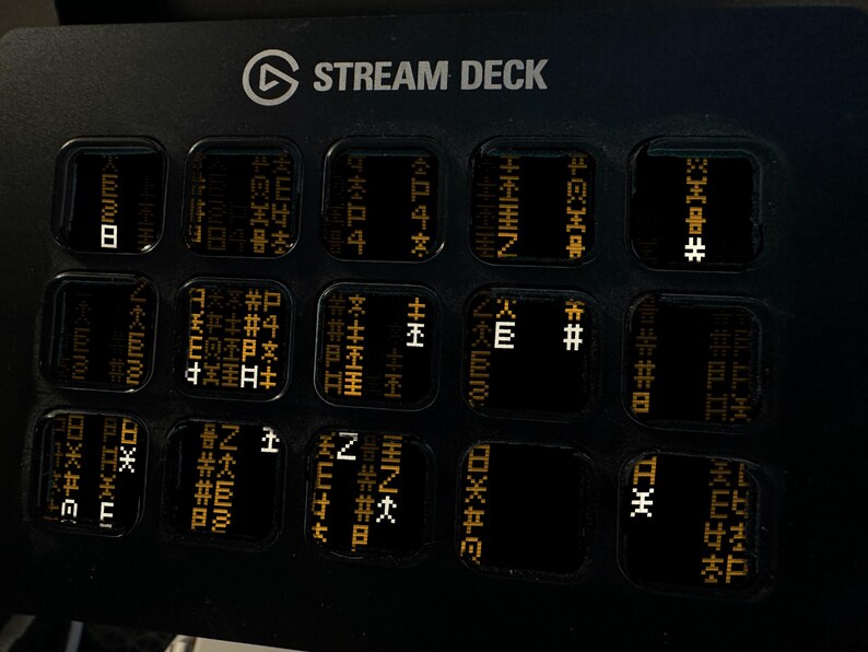 May include: A black Stream Deck control console featuring fifteen square keys arranged in a grid. Each key displays a unique digital interface with orange and white pixelated symbols and characters against a dark background.