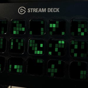 May include: A black Stream Deck control console featuring fifteen square keys arranged in a grid. Each key displays a glowing green digital pattern consisting of small, stacked rectangular blocks against a dark background, with the text STREAM DECK printed in white at the top.