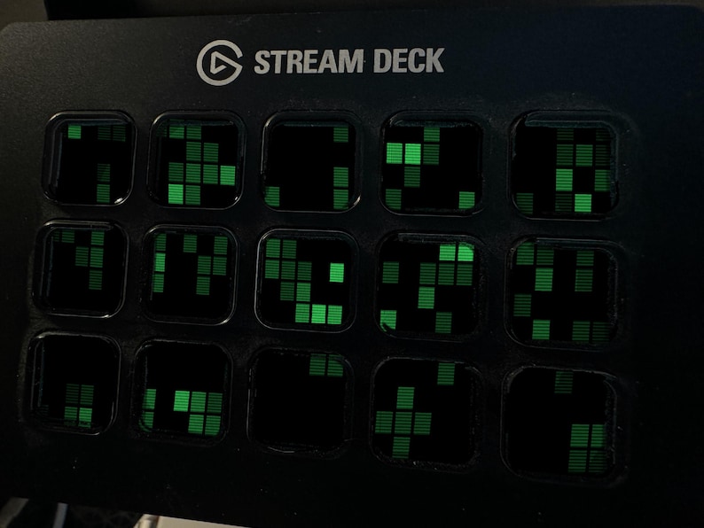 May include: A black Stream Deck control console featuring fifteen square keys arranged in a grid. Each key displays a glowing green digital pattern consisting of small, stacked rectangular blocks against a dark background, with the text STREAM DECK printed in white at the top.
